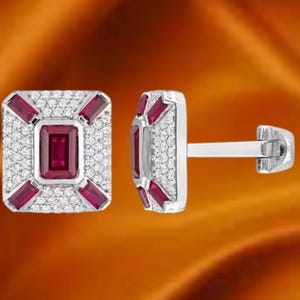 May include: A pair of square cufflinks featuring a central rectangular ruby and smaller ruby accents. The cufflinks are set with small, sparkling white stones in a silver-coloured metal. The background is a warm, orange-toned gradient.