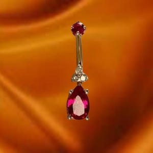 Op de afbeelding: Een zilveren navelpiercing met een druppelvormige, dieprode edelsteen. De ring heeft een kleine, ronde rode edelsteen aan de bovenkant en een cluster van kleine, heldere edelstenen. De achtergrond is een warme, oranje getinte stof.