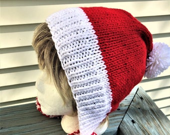 Könnte beinhalten: Ein rot-weißer gestrickter Weihnachtsmannhut mit weißer Krempe und weißem Bommel. Der Hut wird auf einem Schaufensterkopf präsentiert. Der Hut hat eine lange, spitze Form und ist aus weichem, strukturiertem Garn gefertigt. Der Bommel ist groß und flauschig.