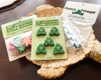 Könnte beinhalten: Eine Sammlung von Bastelartikeln zum Thema Frosch. Dazu gehören Holzknöpfe, eine Holz-Schmuckkette und Schnürsenkel. Die Knöpfe und Schnürsenkel weisen Froschmotive auf. Die Schmuckkette hat Holzblumen und andere Formen.