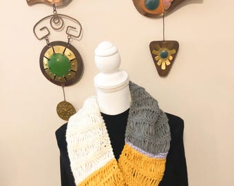 Könnte beinhalten: Handgestrickter Infinity-Schal in Weiß, Gelb und Grau. Der Schal wird auf einem weißen Schaufensterkopf präsentiert und hat ein strukturiertes, offenes Webdesign. Dekorative Metallvögel hängen im Hintergrund.