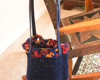 Könnte beinhalten: Eine handgefertigte Tragetasche mit marineblauem Korpus und einem farbenfrohen Oberteil. Der obere Bereich hat rote, orangefarbene und gelbe Garne. Die Tasche hat dunkelblaue Ledergriffe.
