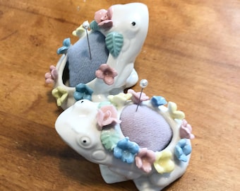 Könnte beinhalten: Zwei weiße, froschförmige Nadelkissen aus Keramik mit pastellfarbenen Blumenakzenten in Rosa, Blau und Gelb. Jeder Frosch hat ein weiches, rundes Samtkissen auf dem Rücken, in das jeweils eine silberfarbene Stecknadel eingesteckt ist.