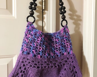Könnte beinhalten: Handgefertigte Häkeltasche in Lila, Blau und Rosa. Die Tasche hat ein einzigartiges Lochmuster und einen ausgestellten Boden. Sie hat einen schwarzen Griff mit Holzperlen und schwarzen Ringen.