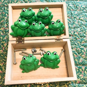 May include: A set of seven green frog-shaped cabinet knobs with white and black eyes, stored in a light-colored wooden box. The set includes several metal bolts and hardware pieces for installation, all resting inside the open wooden container.