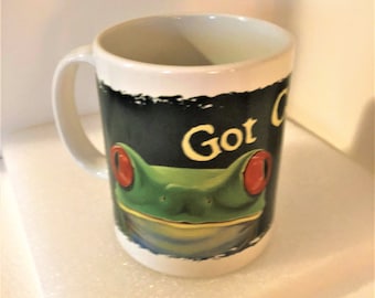 Könnte beinhalten: Weiße Keramiktasse mit einem grünen Froschgesicht-Design und den gelben Worten "Got Coffee". Der Frosch hat rote Augen und ist vor einem schwarzen Hintergrund platziert. Die Tasse hat einen weißen Henkel und Innenraum.