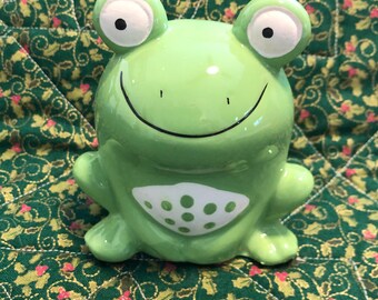 Könnte beinhalten: Eine glänzende Keramik-Froschfigur in Hellgrün. Der Frosch hat große weiße Augen mit schwarzen Pupillen, einen dünnen schwarzen Mundstrich und einen weißen Bauchfleck, der mit mehreren hellgrünen kreisförmigen Punkten versehen ist.