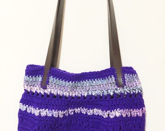 Könnte beinhalten: Handgefertigte, lila gehäkelte Tragetasche mit braunen Ledergriffen. Die Tasche hat horizontale Streifen in verschiedenen Farben, darunter Rosa, Weiß und Blau. Die Tasche hängt an einem Haken.
