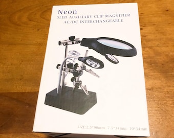 Könnte beinhalten: Eine weiße Box mit dem Text "Neon 5LED AUXILIARY CLIP MAGNIFIER AC/DC INTERCHANGEABLE" zeigt eine Lupe mit LED-Leuchten und verstellbaren Clips. Die Box gibt auch die Größe als 2,5*90mm, 7,5*34mm und 10*34mm an.