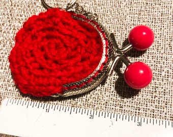 Könnte beinhalten: Ein rotes gehäkeltes Münzfach mit silbernem Verschluss und Schlüsselring. Die Geldbörse hat ein rundes, strukturiertes Design und ist mit zwei roten, runden Perlen akzentuiert. Ein bezauberndes Accessoire.