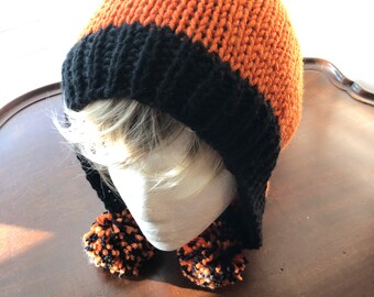 Könnte beinhalten: Eine orange-schwarze Strickmütze mit Ohrenklappen und zwei Bommeln. Die Mütze ist hauptsächlich orange mit einem schwarzen Band am unteren Rand. Die Bommeln sind schwarz und orange. Die Mütze wird auf einem Schaufensterkopf präsentiert.