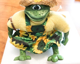 Könnte beinhalten: Eine grüne Froschfigur mit einem Strohhut, der mit einer gelben Blume verziert ist, einem gelben kurzärmeligen Strickoberteil und einer grünen Schürze mit Sonnenblumenmuster. Der Frosch hat eine texturierte Haut, große Augen und einen breiten Mund.