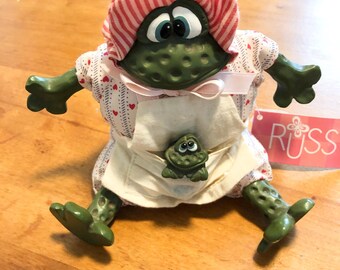 Könnte beinhalten: Eine grüne Froschfigur mit einer rot-weiß gestreiften Haube und einem weißen Kleid mit rotem Herzmuster. Der Frosch trägt eine weiße Schürze mit einer kleinen Froschfigur in der Tasche und ein rosa Etikett der Marke Russ an der Seite.