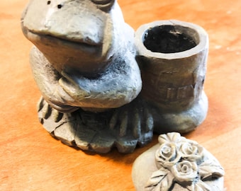 Könnte beinhalten: Ein grauer, froschförmiger Pflanztopf mit einem kleinen, runden Deckel mit Rosenmuster. Der Pflanztopf hat eine zylindrische Öffnung für Pflanzen und besteht aus einem steinartigen Material. Der Frosch sitzt mit verschränkten Armen.