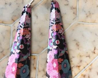 Könnte beinhalten: Ein Paar lange, trapezförmige Ohrringe mit floralem Muster. Die Ohrringe haben einen schwarzen Hintergrund mit rosa, blauen und orangefarbenen Blumen. Die Ohrringe haben silberfarbene Ohrhaken.