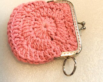 Könnte beinhalten: Ein kleines, rundes, gehäkeltes Münztäschchen in Rosa mit einem silberfarbenen Metall-Bügelverschluss mit Kugelverschluss. Ein runder silberner Metallring ist an der Seite des Rahmens befestigt, sodass es als Schlüsselanhänger verwendet werden kann.