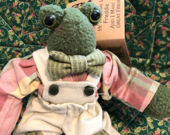 Könnte beinhalten: Ein Frosch-Stofftier mit dunkelgrüner, strukturierter Haut und großen schwarzen Augen. Er trägt eine grün karierte Fliege, ein langärmeliges Hemd mit rosa-gelbem Karomuster und eine cremefarbene Latzhose mit zwei schwarzen Knöpfen und Flicken.