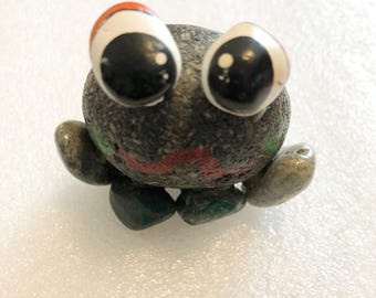 Könnte beinhalten: Eine kleine Kreaturenfigur aus glatten grauen Flusskieselsteinen. Sie hat zwei große, runde, weiß bemalte Augen mit schwarzen Pupillen und einen kleinen rot bemalten Mund. Der Körper besteht aus einem zentralen Stein mit kleineren Steinen als Gliedmaßen.