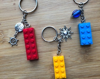 Könnte beinhalten: Drei Schlüsselanhänger mit verschiedenen farbigen Lego-Stein-Anhängern. Der erste Schlüsselanhänger hat einen roten Lego-Stein-Anhänger mit einem silbernen nautischen Rad-Anhänger. Der zweite Schlüsselanhänger hat einen gelben Lego-Stein-Anhänger mit einem silbernen Sonnen-Anhänger. Der dritte Schlüsselanhänger hat einen blauen Lego-Stein-Anhänger mit einem silbernen Anhänger und einer blauen Glasperle.
