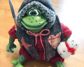 Könnte beinhalten: Eine grüne Froschfigur, die eine dunkelgraue Kapuzenweste über einem roten Outfit trägt. Die Weste ist mit einem roten Stern und einem karierten Herzaufnäher verziert. Der Frosch hält einen kleinen Holzstab und einen weißen Schneemann.