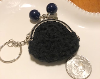 Könnte beinhalten: Eine kleine schwarze gehäkelte Münzbörse als Schlüsselanhänger mit einem silberfarbenen Metall-Kussverschluss, der mit zwei dunkelblauen, kugelförmigen Perlen verziert ist. Eine US-Vierteldollarmünze liegt daneben als Größenvergleich.