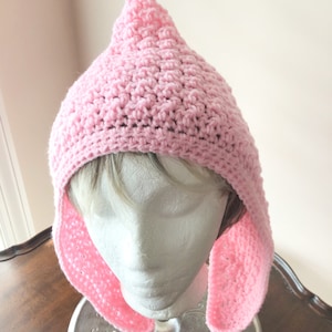 May include: A light pink crocheted hat with a pointed top and ear flaps. The hat is displayed on a mannequin head. The hat is made of a soft, textured yarn, and the ear flaps extend down the sides.