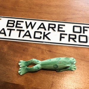 May include: A rectangular white sign with a black border and the black text BEWARE OF ATTACK FROG, accompanied by a small, hand-carved wooden frog figurine painted in light green with darker green spots, positioned below the sign.