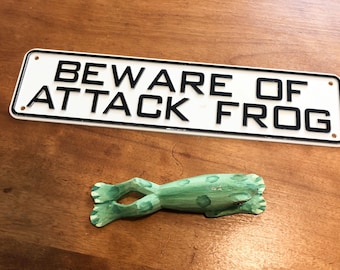 Könnte beinhalten: Ein rechteckiges weißes Schild mit schwarzem Rand und dem schwarzen Text BEWARE OF ATTACK FROG, begleitet von einer kleinen, handgeschnitzten Froschfigur aus Holz, die hellgrün mit dunkelgrünen Flecken bemalt ist und unter dem Schild liegt.