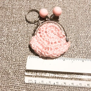 May include: A small, pink crocheted coin purse with a metal clasp and a key ring. Two pink beads are attached to the top of the purse. The purse is approximately 2 inches wide.