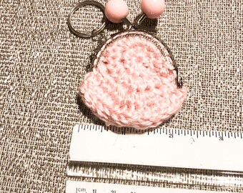 Könnte beinhalten: Ein kleiner, rosa gehäkelter Münzbeutel mit Metallverschluss und Schlüsselring. Zwei rosa Perlen sind oben am Beutel befestigt. Der Beutel ist etwa 5 cm breit.