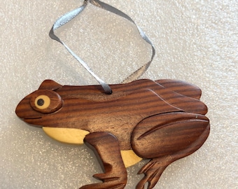 Könnte beinhalten: Ein hölzernes Frosch-Ornament mit dunkelbrauner Holzmaserung und hellen cremefarbenen Akzenten an Auge, Kehle und Bauch. Der Frosch ist in sitzender Position geformt, mit geschnitzten Details an Beinen und Rücken, und enthält ein dünnes silbernes Band.