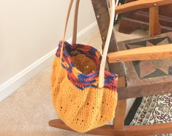 Könnte beinhalten: Eine gelbe Häkeltasche mit hellbraunen Ledergriffen. Die Oberseite der Tasche hat ein farbenfrohes Muster aus Rot, Blau und Gelb. Die Tasche hängt an einem hölzernen Schaukelstuhl.