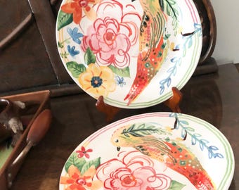 Könnte beinhalten: Zwei dekorative Keramikplatten mit Vogel- und Blumendesign. Die Teller haben einen weißen Hintergrund mit einer farbenfrohen Illustration eines Vogels, rosa Rosen, orangefarbenen Blumen und blauen Akzenten. Die Teller stehen auf Holzständern.