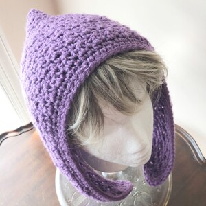May include: A handmade, purple crochet hat with a pointed top and long, ear-like flaps. The hat is made of a textured yarn and appears to be designed to fit snugly. The hat is displayed on a mannequin head.