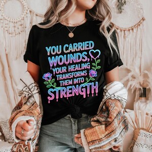 Peut inclure: T-shirt noir avec l'inscription « YOU CARRIED WOUNDS, YOUR HEALING TRANSFORMS THEM INTO STRENGTH » en dégradé néon, ornée d'un cœur et de motifs floraux. Le t-shirt est porté avec un jean et une veste à motifs.