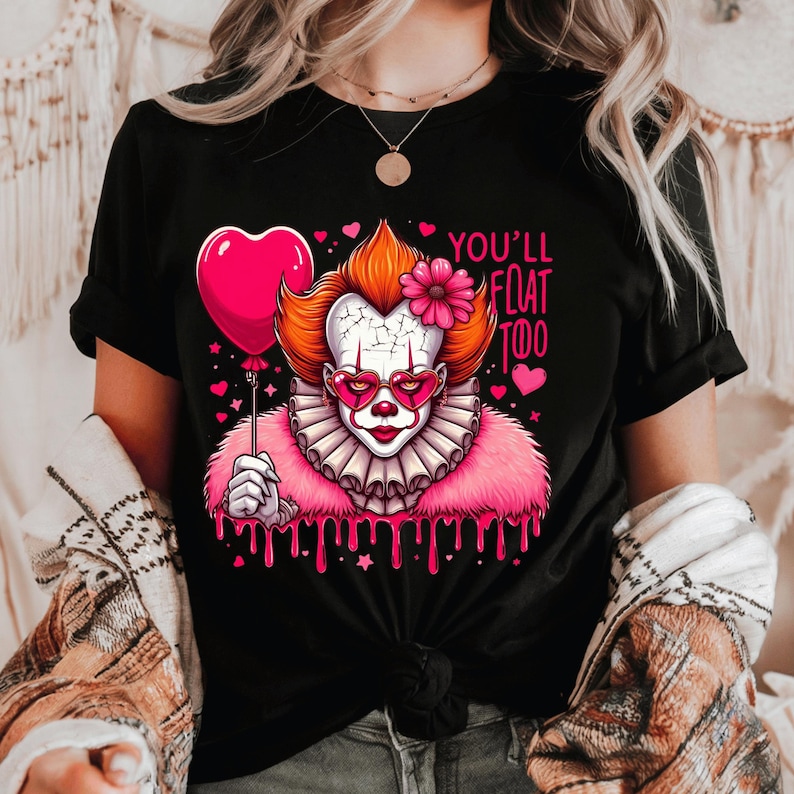 May include: Black t-shirt featuring a colorful graphic of a clown with heart-shaped glasses and a pink balloon. The clown has orange hair, a pink flower, and the text "YOU'LL FLOAT TOO". The design includes pink hearts and dripping pink accents.