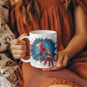 May include: White ceramic mug featuring a 3D graphic of Spider-Man breaking through a wall. The superhero is depicted in his classic red and blue suit, with a web and blue background.