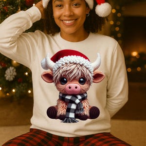 Peut inclure: Sweat-shirt crème avec une vache des Highlands crochetée portant un bonnet de Père Noël et une écharpe à carreaux noirs et blancs. La vache a de grands yeux expressifs et des cornes blanches. Collection sur le thème de Noël.