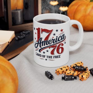 Peut inclure: Un mug en céramique blanc orné d'un motif graphique rouge, blanc et bleu avec le texte 17 America 76 et Land of the Free. Le design comprend des étoiles et des textures usées, avec une anse sur le côté droit et un liquide sombre à l'intérieur.