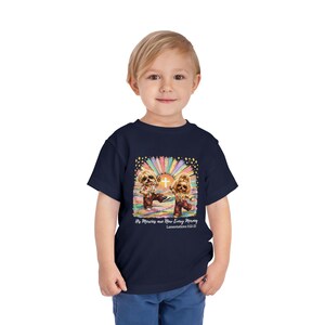 Peut inclure: Un t-shirt bleu marine à manches courtes avec un motif coloré représentant deux chiens vêtus de vêtements à motifs devant une croix lumineuse et un soleil. Le texte indique "His Mercies are New Every Morning" et "Lamentations 3:22-23".