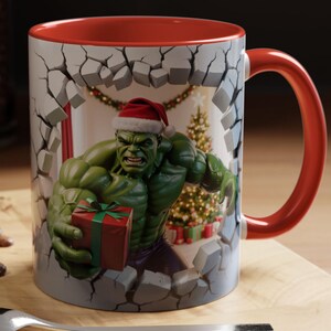 Peut inclure: Mug en c&eacute;ramique avec le Hulk portant un bonnet de P&egrave;re No&euml;l, per&ccedil;ant un mur. L'int&eacute;rieur et la anse sont rouges. Le Hulk tient un cadeau, avec un sapin de No&euml;l et des d&eacute;corations en arri&egrave;re-plan.