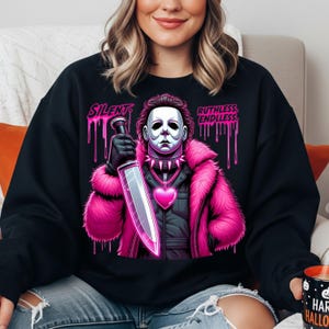 May include: Black sweatshirt featuring a graphic of a masked figure in a pink fur coat, holding a large knife. The design includes the words "SILENT" and "RUTHLESS: ENDLESS" in pink dripping font. A heart-shaped pendant is visible.