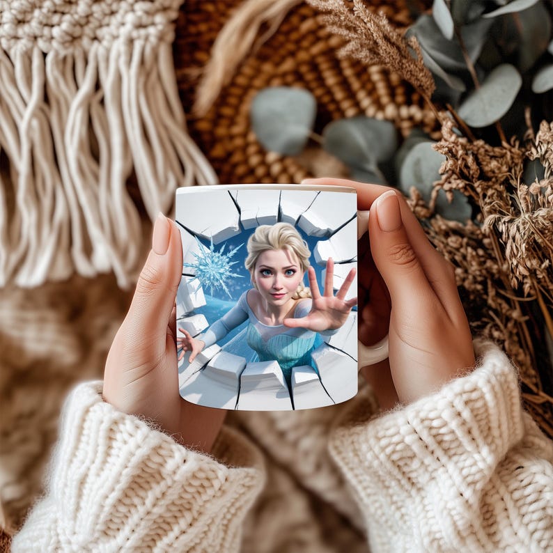 May include: White ceramic mug featuring a graphic of a woman in a blue dress reaching out from a cracked wall. The mug has a glossy finish and is held by a person wearing a cream-colored sweater. The background includes dried plants.