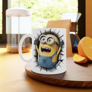 Puede incluir: Taza de cerámica blanca con un gráfico 3D de un personaje Minion rompiendo una pared agrietada. El Minion es amarillo con guantes y gafas negros, vistiendo un peto azul. La taza tiene un asa blanca y está sobre una superficie de madera.