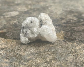 May include: A cluster of small, light-colored crystals with a rough, textured surface. The crystals have a slightly translucent appearance and are clustered together. The overall color is a pale white with some darker specks.