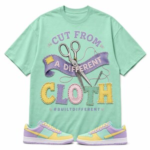 May include: A mint green short-sleeve t-shirt featuring a graphic of silver scissors, a needle, and a purple banner with the text CUT FROM A DIFFERENT CLOTH, along with the word CLOTH in colorful stitched letters and #BUILTDIFFERENT below.