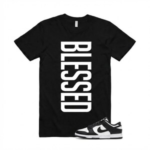Pu&ograve; includere: T-shirt nero con la scritta "BLESSED" in grandi lettere maiuscole bianche. Un paio di scarpe da ginnastica bianche e nere &egrave; posizionato nella parte inferiore della maglietta. Le scarpe da ginnastica hanno una suola bianca e un logo Nike nero.