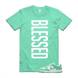 Pu&ograve; includere: Una maglietta verde menta con la parola "BLESSED" stampata verticalmente in bianco. Un paio di scarpe da ginnastica verde menta e bianche sono in primo piano, a complemento della combinazione di colori della maglietta. Le scarpe da ginnastica hanno un logo bianco.