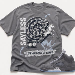 May include: A charcoal gray t-shirt featuring a graphic of a circular chessboard with a knight piece, a chess clock, and a crown. The text reads SAYLESS and REAL ONES MOVE IN SILENCE. A matching charcoal gray high-top sneaker is positioned at the bottom right.