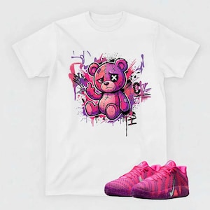 May include: White t-shirt featuring a cartoon teddy bear graphic in shades of pink and purple, giving the middle finger. Below the shirt are pink and purple athletic shoes with a textured design and a Nike swoosh.